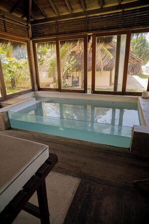 Ein von Glas umschlossener Swimmingpool mit Holzterrasse und einem Tisch in tropischer Umgebung.