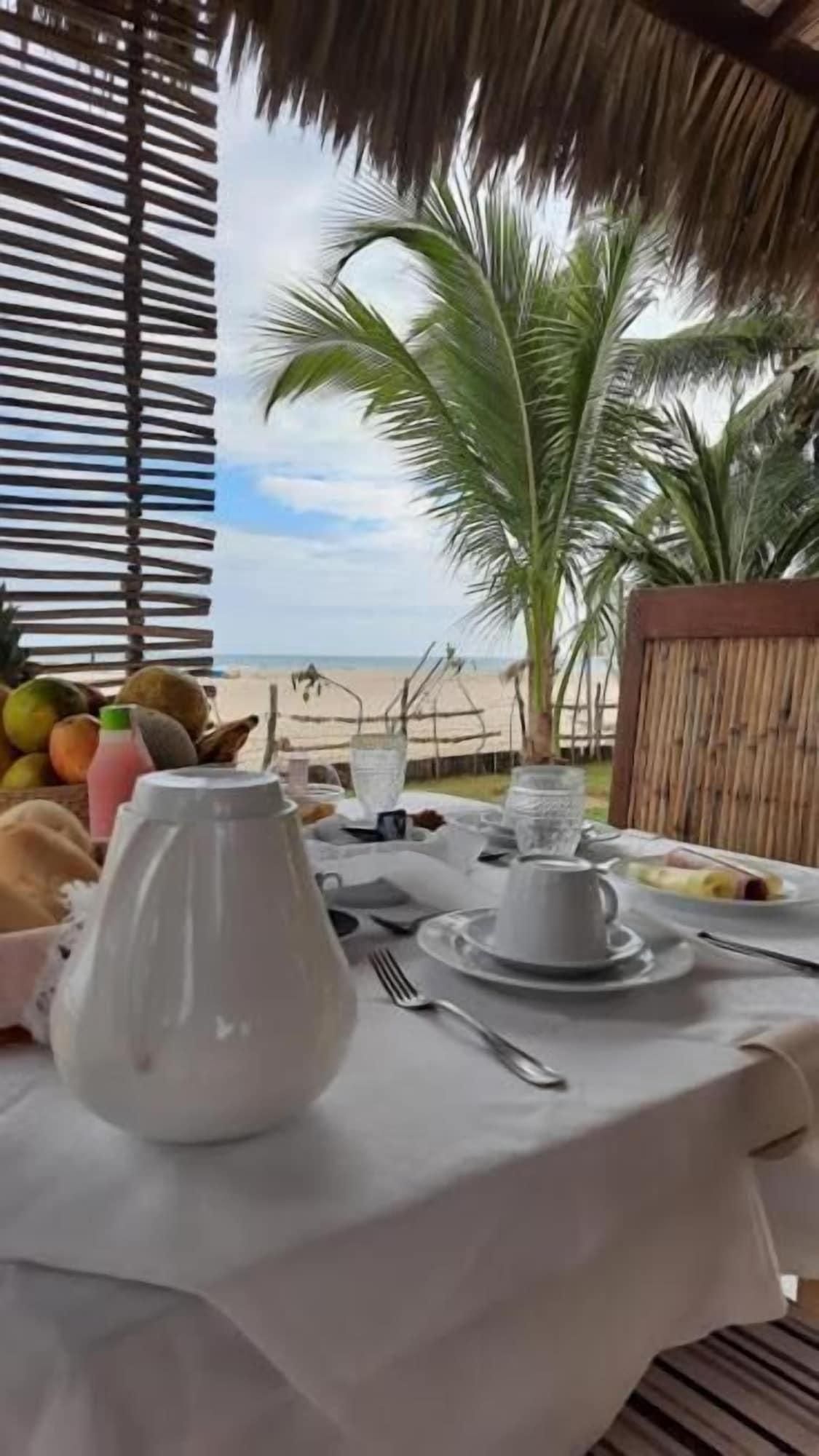 Mesa posta para café da manhã com frutas, uma jarra e xícaras, com vista para uma praia com palmeiras.