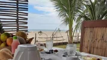 Mesa posta para café da manhã com frutas, uma jarra e xícaras, com vista para uma praia com palmeiras.