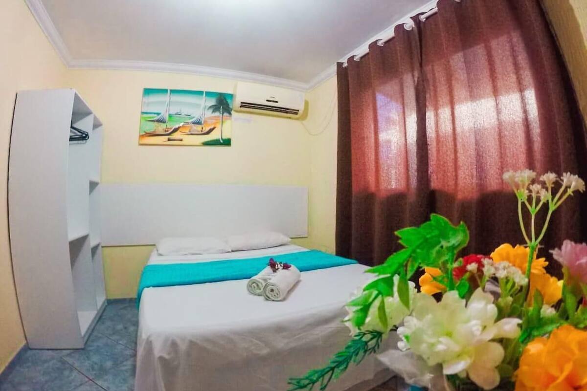 A hotel room with a bed, a painting on the wall, an air conditioner, a curtain, and a vase of flowers.