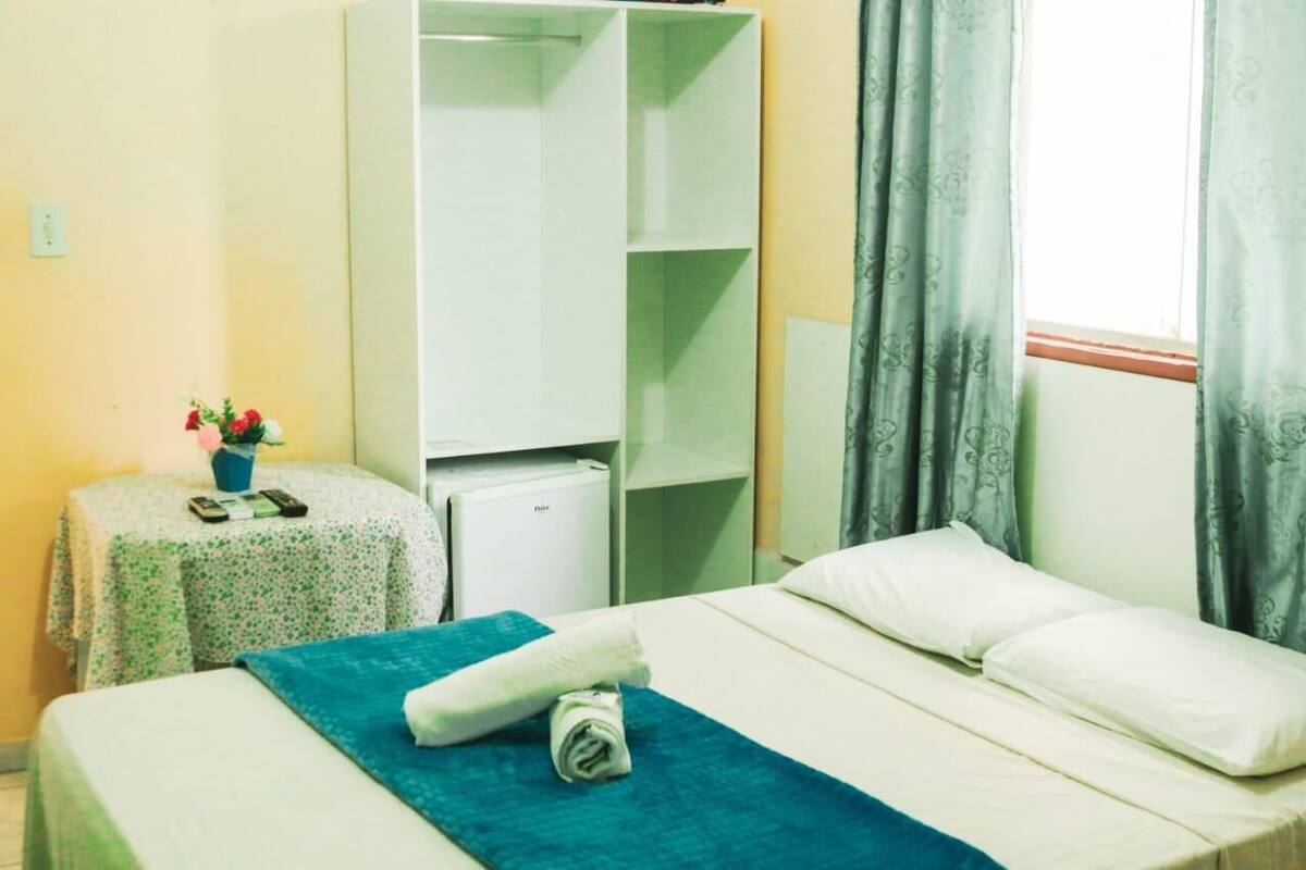 A hotel room with a bed, a small table with a floral tablecloth, a potted plant, a white wardrobe, and a window with curtains.