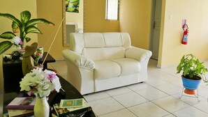 Una sala de estar con un sofá blanco, una mesa de centro con libros y un jarrón de flores, una planta en maceta y un extintor en la pared.