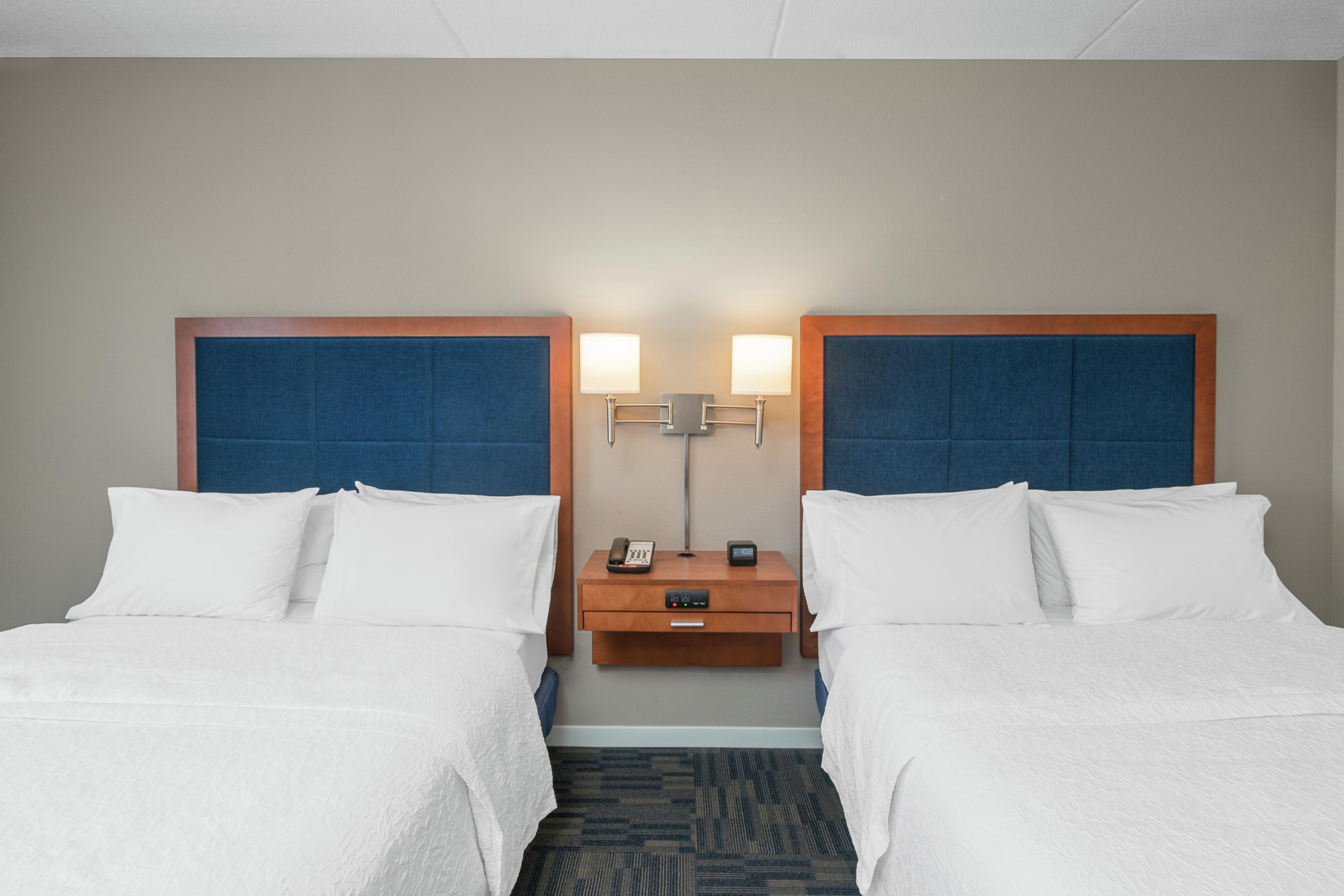 A hotel room with two beds, a nightstand with a lamp, a phone, and a clock.