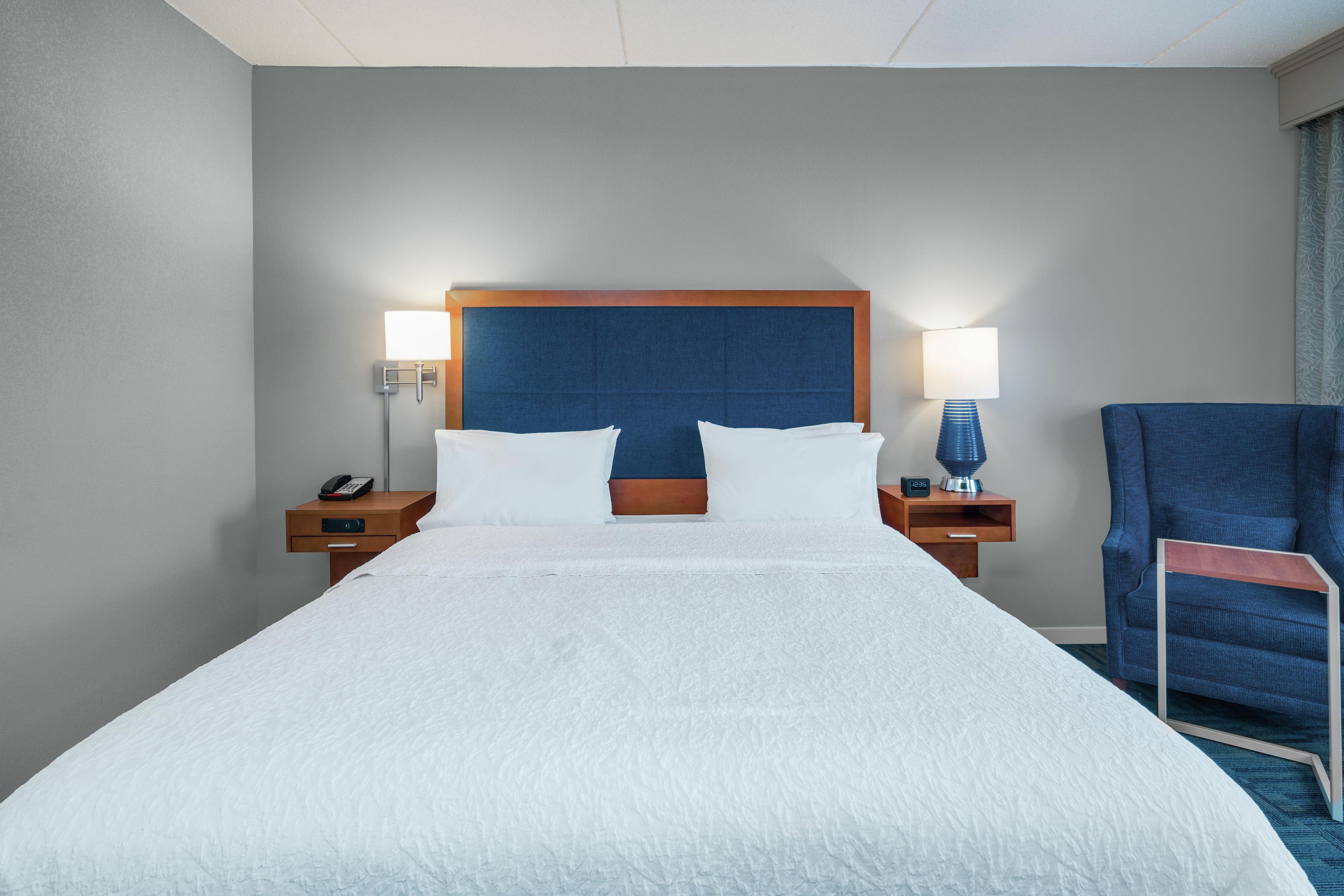 A hotel room with a large bed, two bedside tables, a chair, and a nightstand with a lamp.