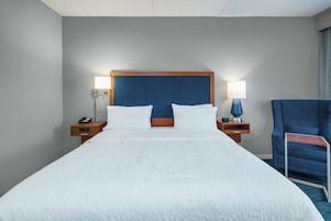 A hotel room with a large bed, two bedside tables, a chair, and a nightstand with a lamp.