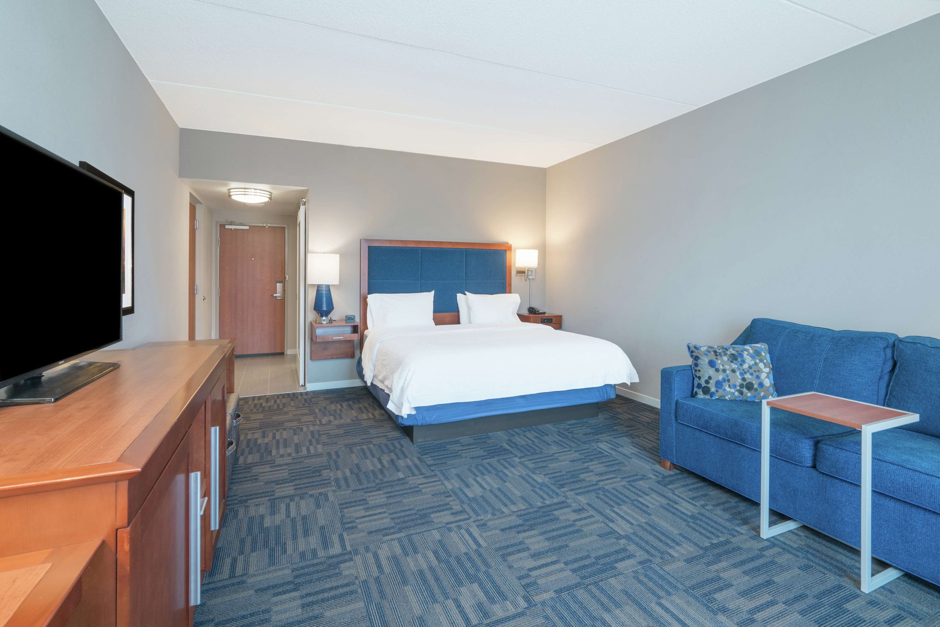 A hotel room with a bed, a blue sofa, a TV, a desk, and a lamp.