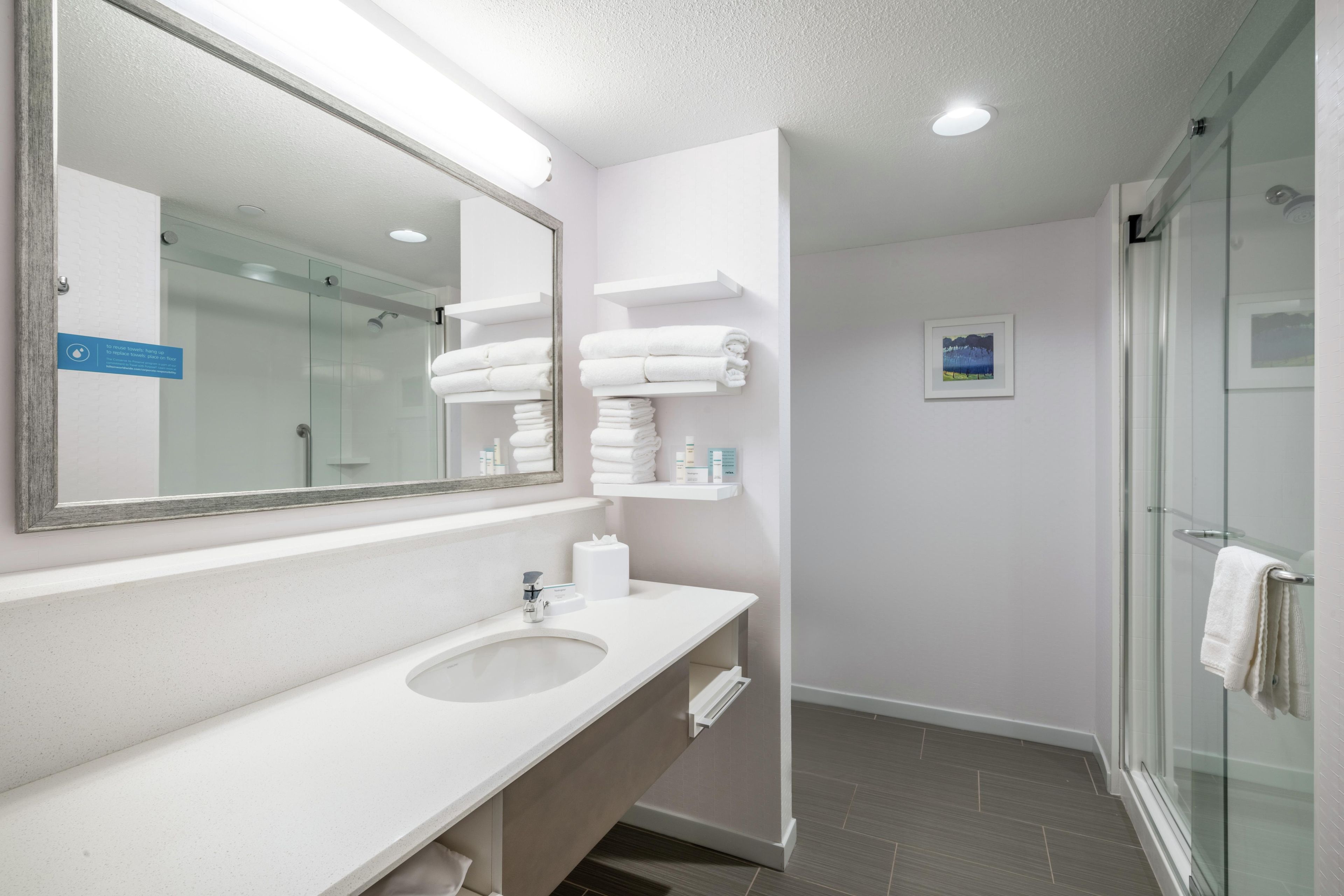 A modern bathroom with a large mirror, a sink, a glass shower enclosure, and neatly folded towels on a shelf.