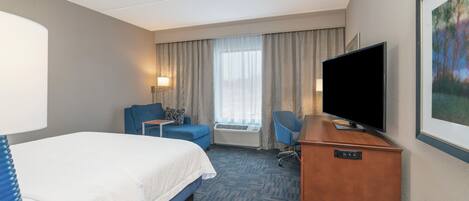 Una habitación de hotel con cama, escritorio, silla, televisor y ventana con cortinas.