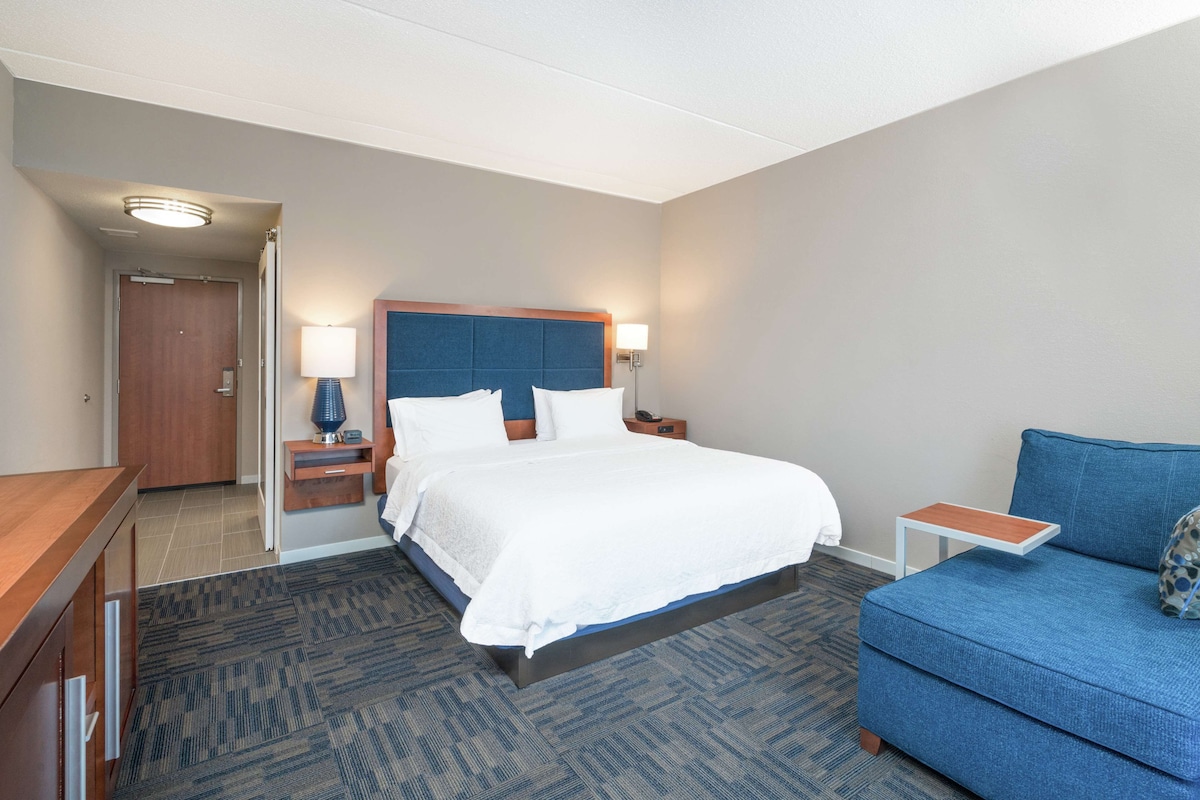 A hotel room with a bed, a blue sofa, a nightstand with a lamp, and a wooden door.
