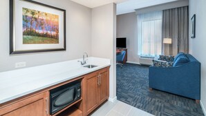 A hotel room with a kitchenette, a living area with a sofa and a TV, and a large window with curtains.