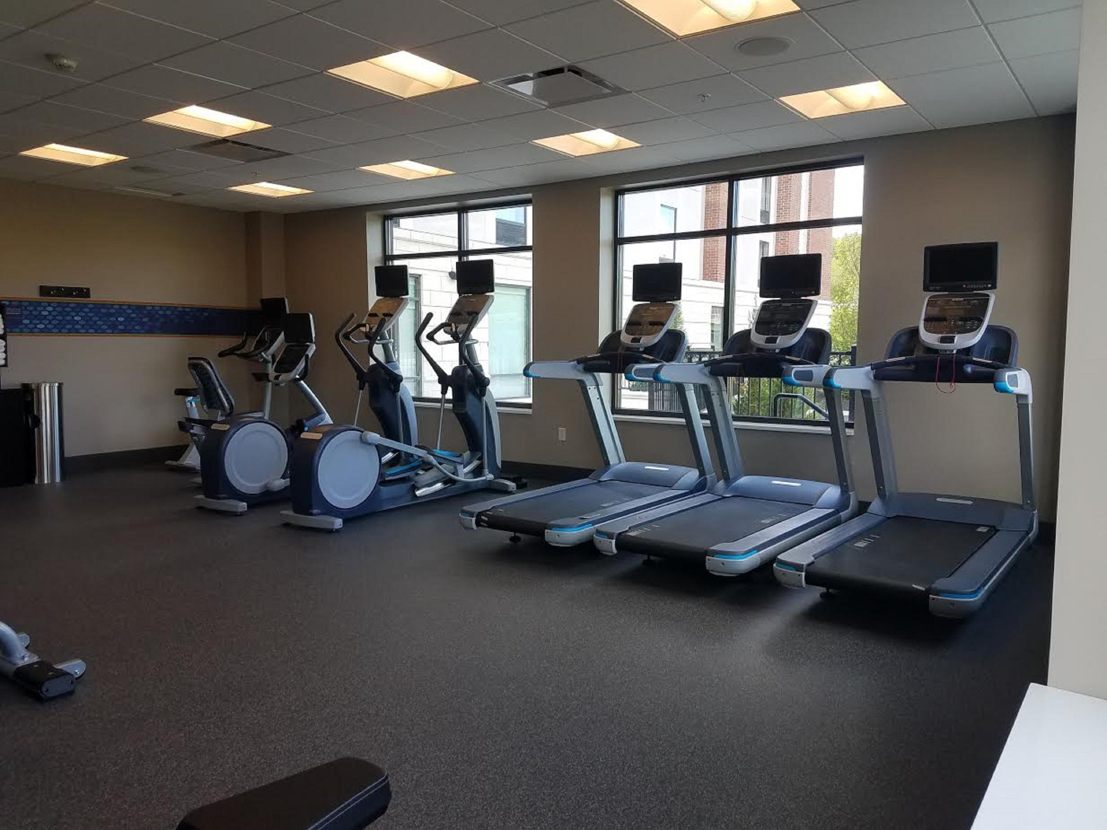 A gym with treadmills, elliptical machines, and a large window with a view of buildings.