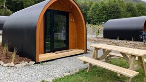 Una estructura de madera semicircular moderna con puerta de vidrio, rodeada de grava y una mesa de picnic.
