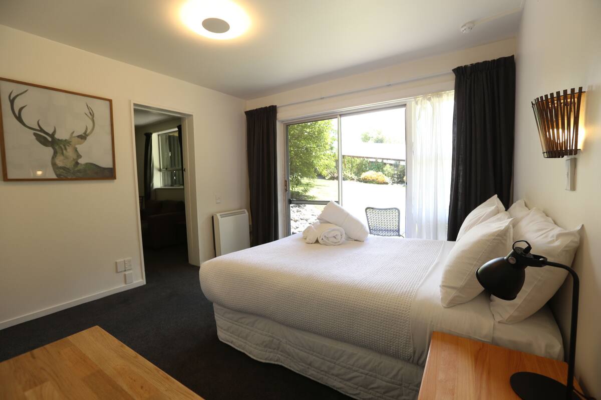 A hotel room with a large bed, a bedside table, a lamp, a window with curtains, a door leading to another room, and a framed deer head artwork on the wall.