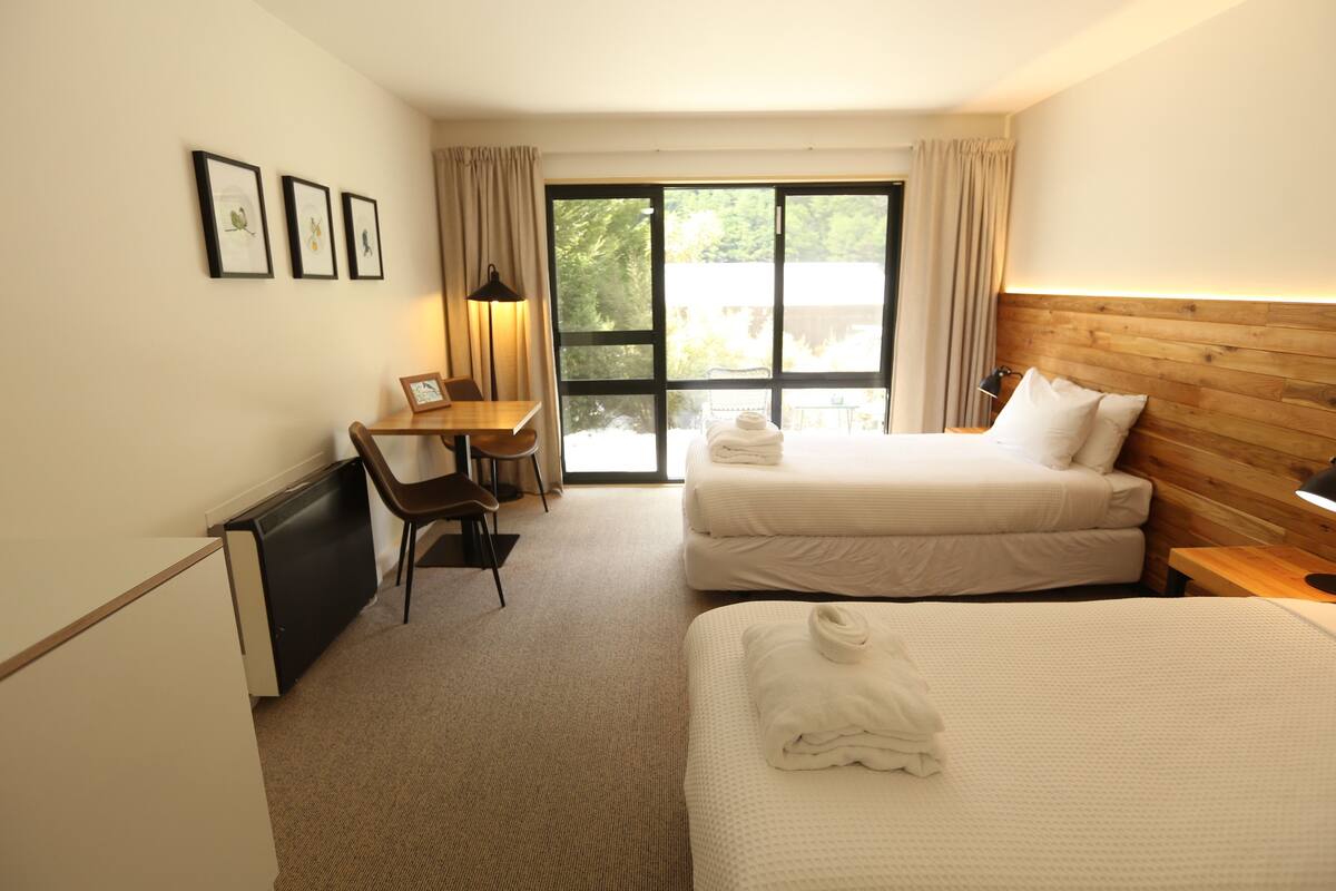 A hotel room with two beds, a desk, a chair, and a large window with curtains.