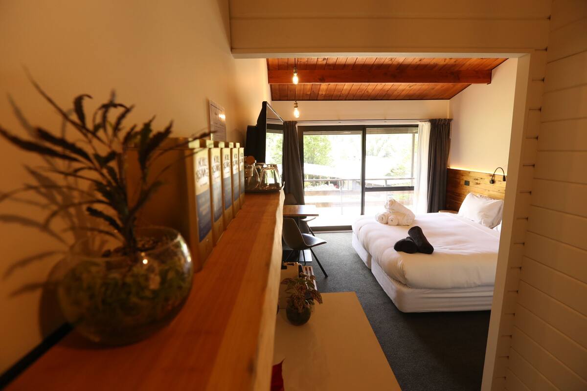 A hotel room with a bed, a wooden desk, a vase with plants, and a large window.