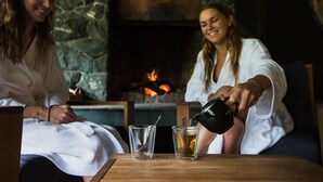 Dos personas sentadas a una mesa con una chimenea de fondo. Una persona está sirviendo una bebida en un vaso.