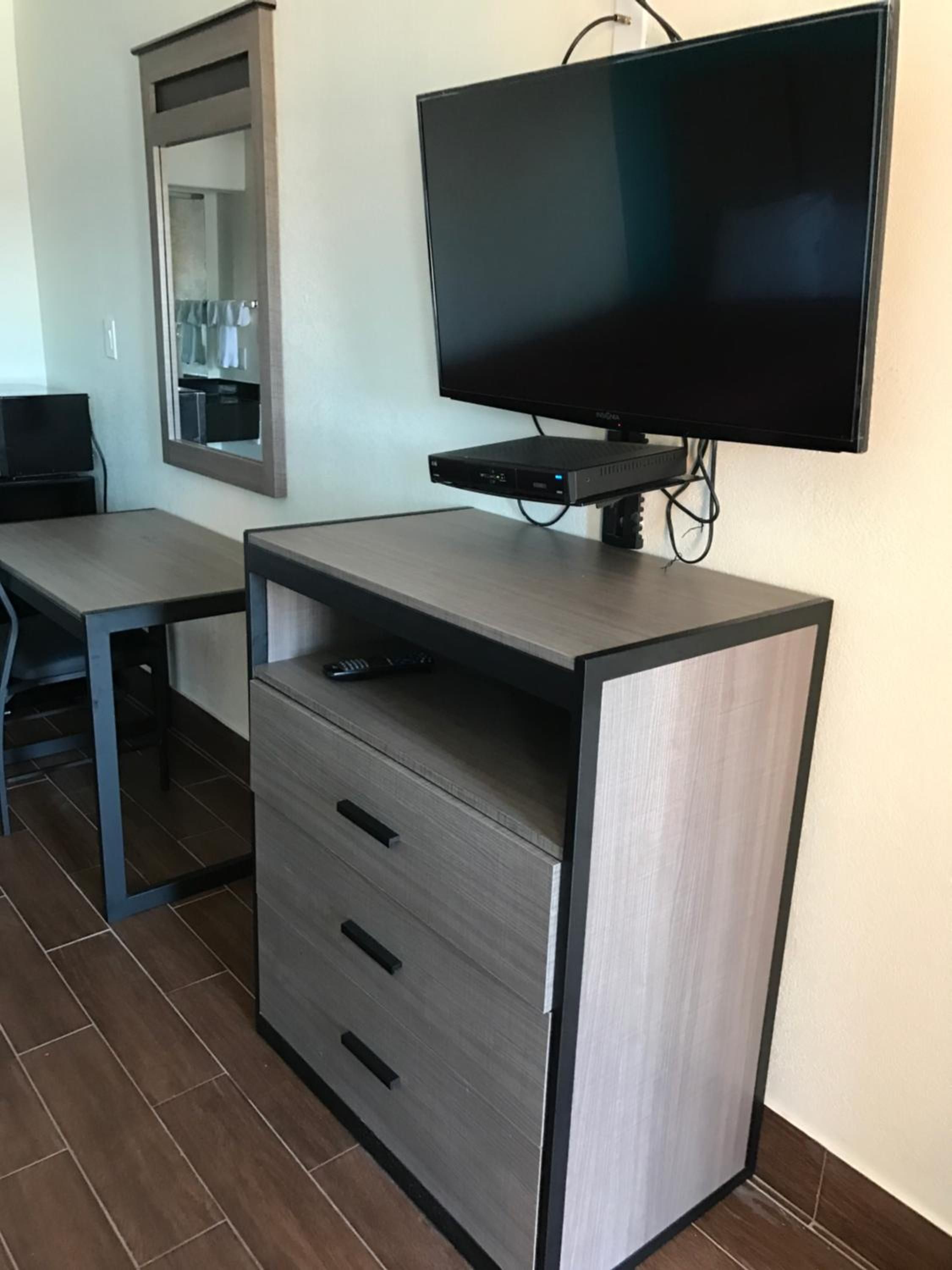 A room with a flat-screen TV mounted on the wall, a desk with a chair, and a cabinet with drawers.