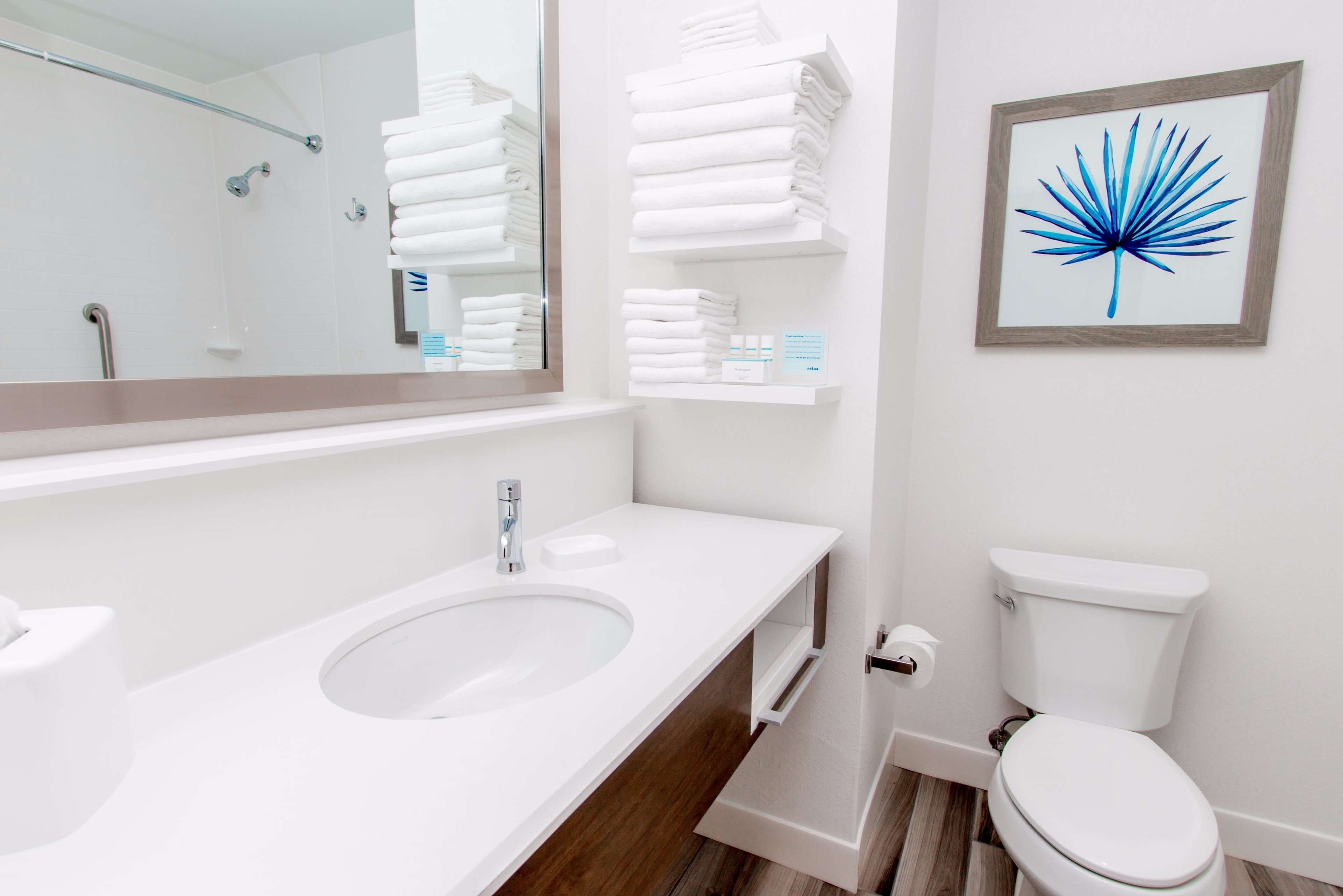 A modern bathroom with a white sink, mirror, and toilet. There are white towels stacked on a shelf and a framed artwork on the wall.