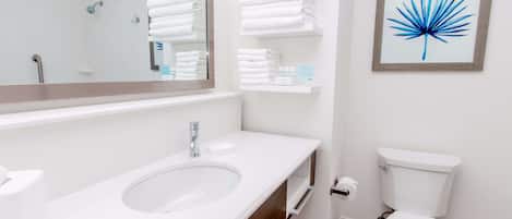 A modern bathroom with a white sink, mirror, and toilet. There are white towels stacked on a shelf and a framed artwork on the wall.