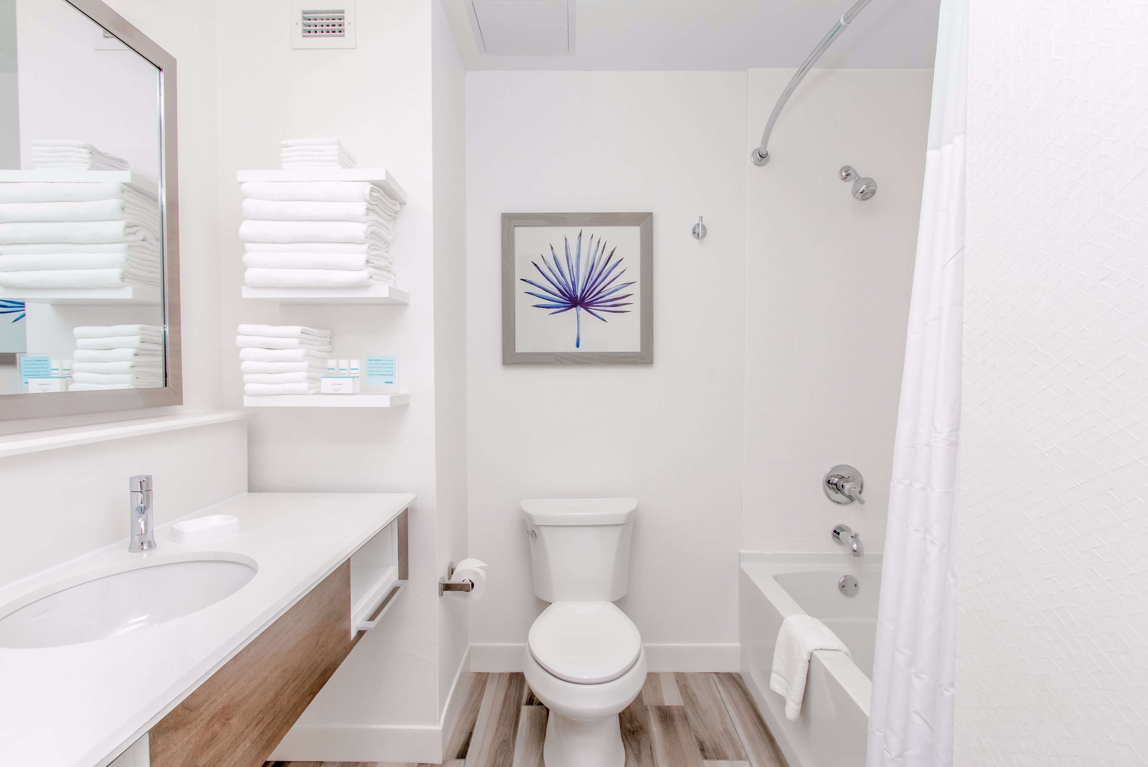 Une salle de bain moderne avec des toilettes, un lavabo et une baignoire. Des serviettes sont pliées et empilées sur des étagères, et un tableau encadré est accroché au mur.