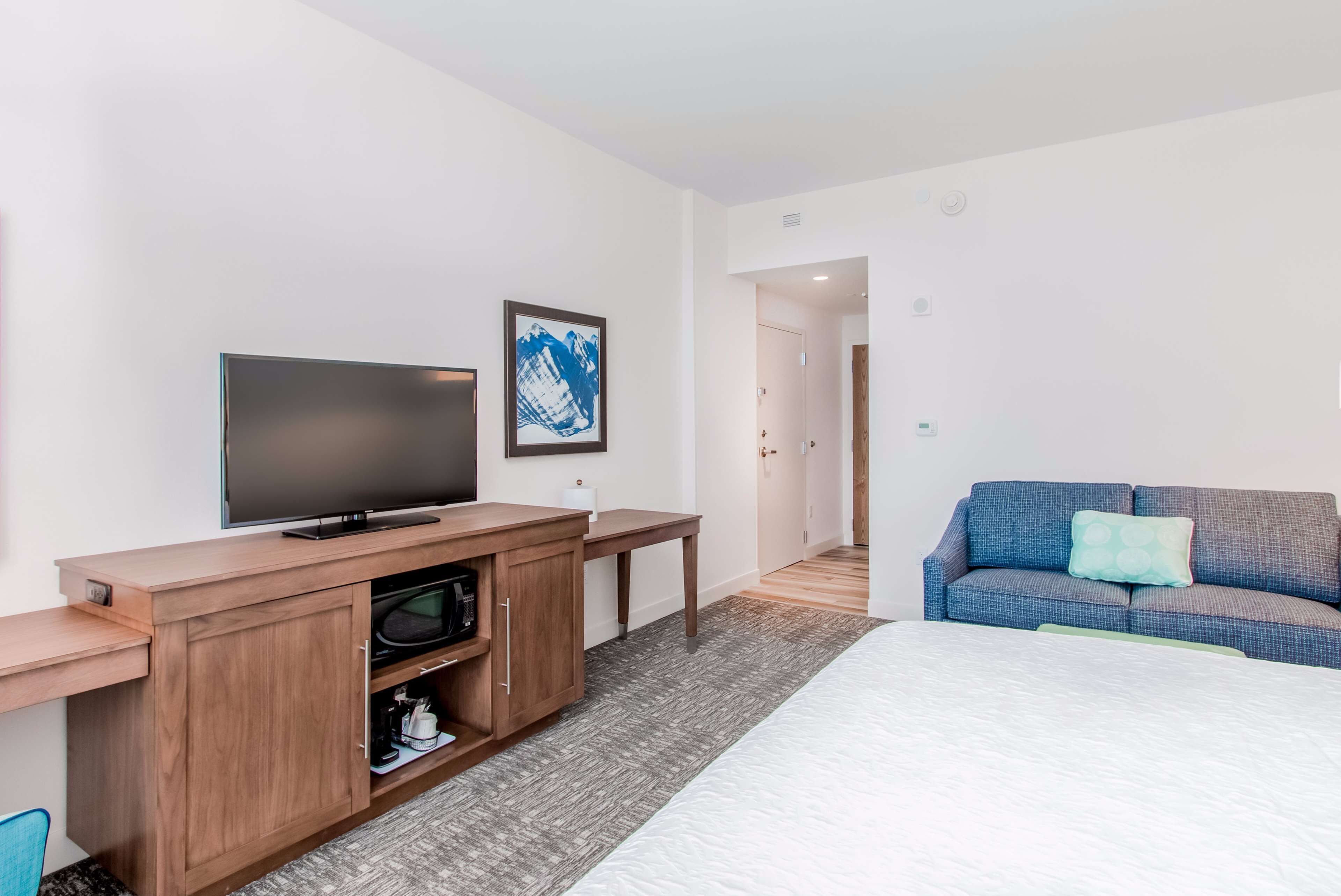 A modern hotel room with a flat-screen TV, a wooden entertainment unit, a bed with a white mattress, a blue sofa with a green pillow, and a framed abstract painting on the wall.