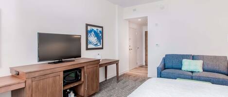 Une chambre d’hôtel moderne équipée d’un téléviseur à écran plat, d’un meuble de rangement en bois, d’un lit avec un matelas blanc, d’un canapé bleu avec un coussin vert et d’un tableau abstrait encadré accroché au mur.