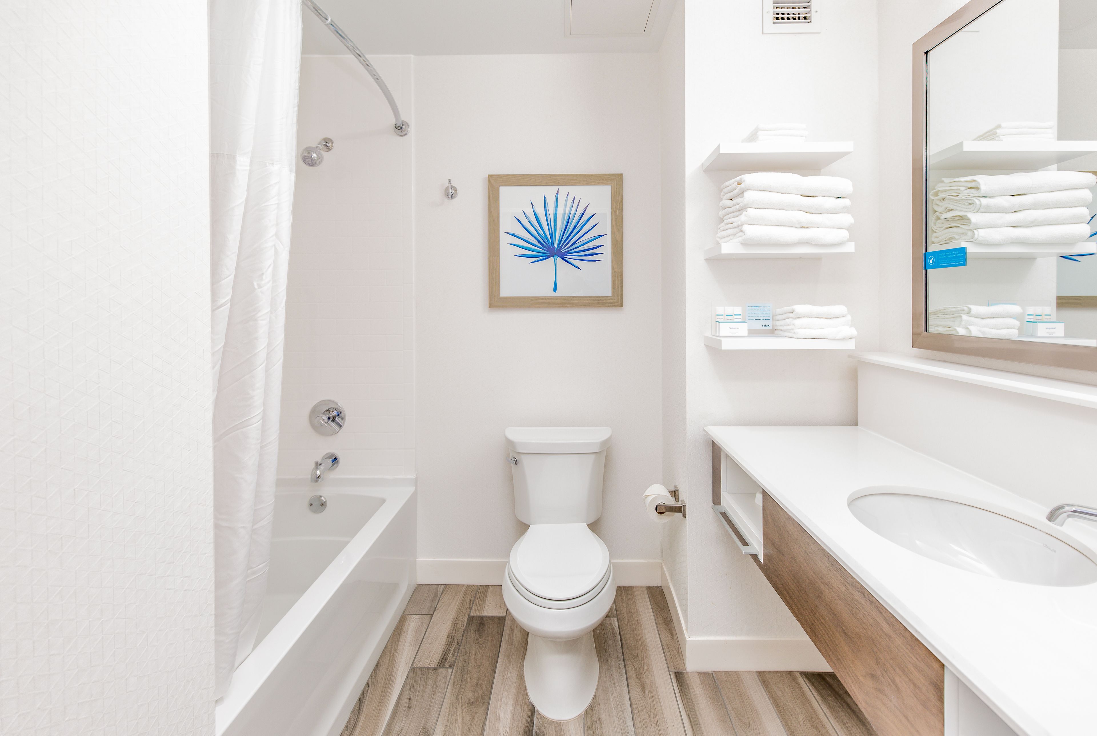A modern bathroom with a white toilet, a bathtub, and a sink. There is a framed artwork on the wall and a towel rack with folded towels.