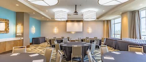 A conference room with round tables, chairs, a projector screen, and a large window with curtains.