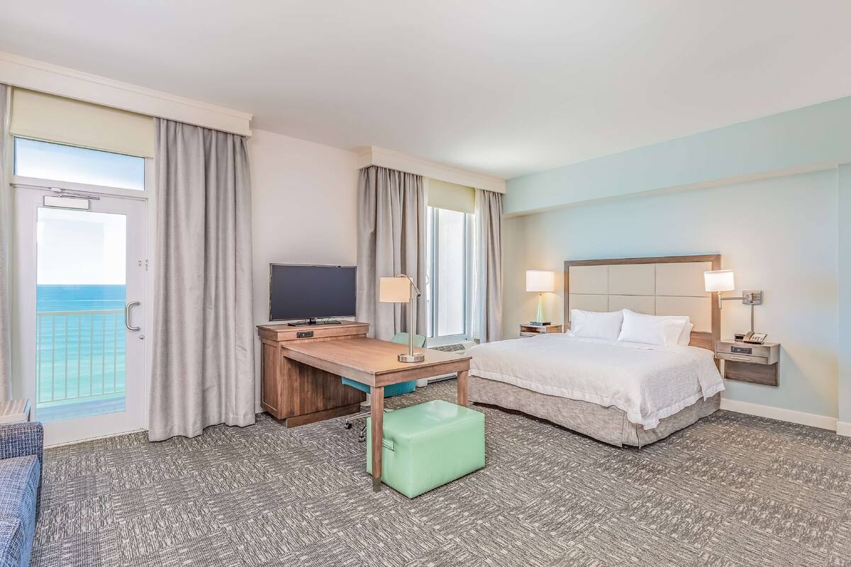 Een hotelkamer met een bed, bureau, stoel en uitzicht op de oceaan door een schuifdeur.