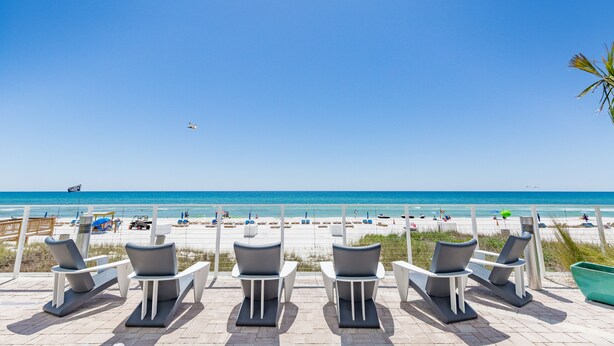 Un salón junto a la playa con sillas orientadas al océano, cielo despejado y un avión a lo lejos.