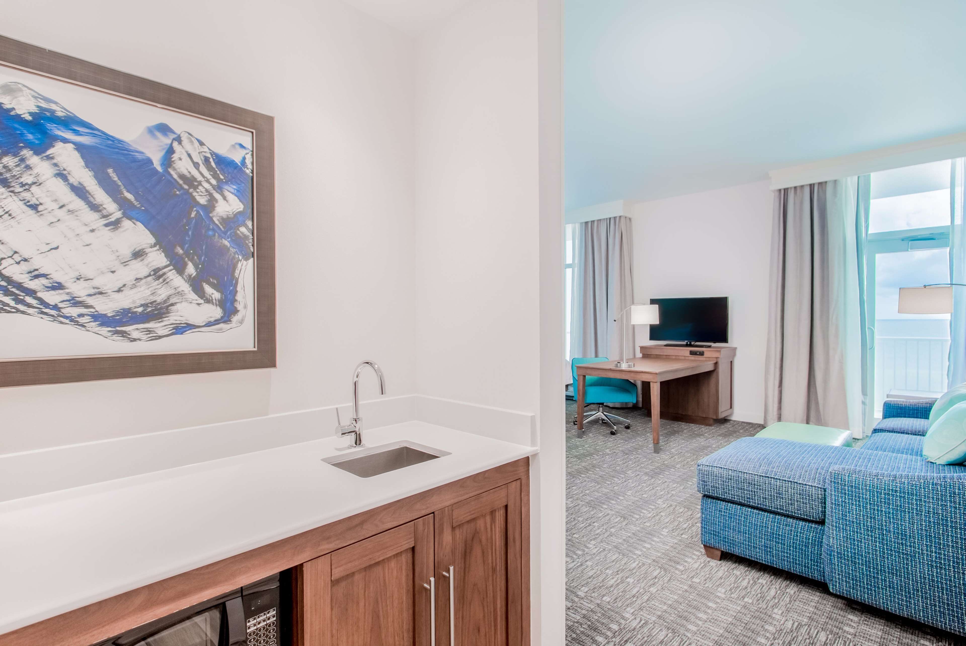 A hotel room with a white sink, a framed abstract painting, a desk with a TV, and a blue sofa.