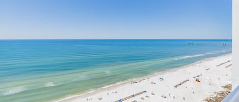 A beach with rows of sun loungers and umbrellas, a pier in the distance, and a clear blue sky.