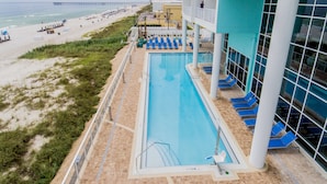 Un área de piscina con vistas a la playa y al océano, un muelle a lo lejos, y un edificio con balcones que dan a la piscina.