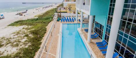 A pool area with a view of the beach and ocean, a pier in the distance, and a building with balconies overlooking the pool.