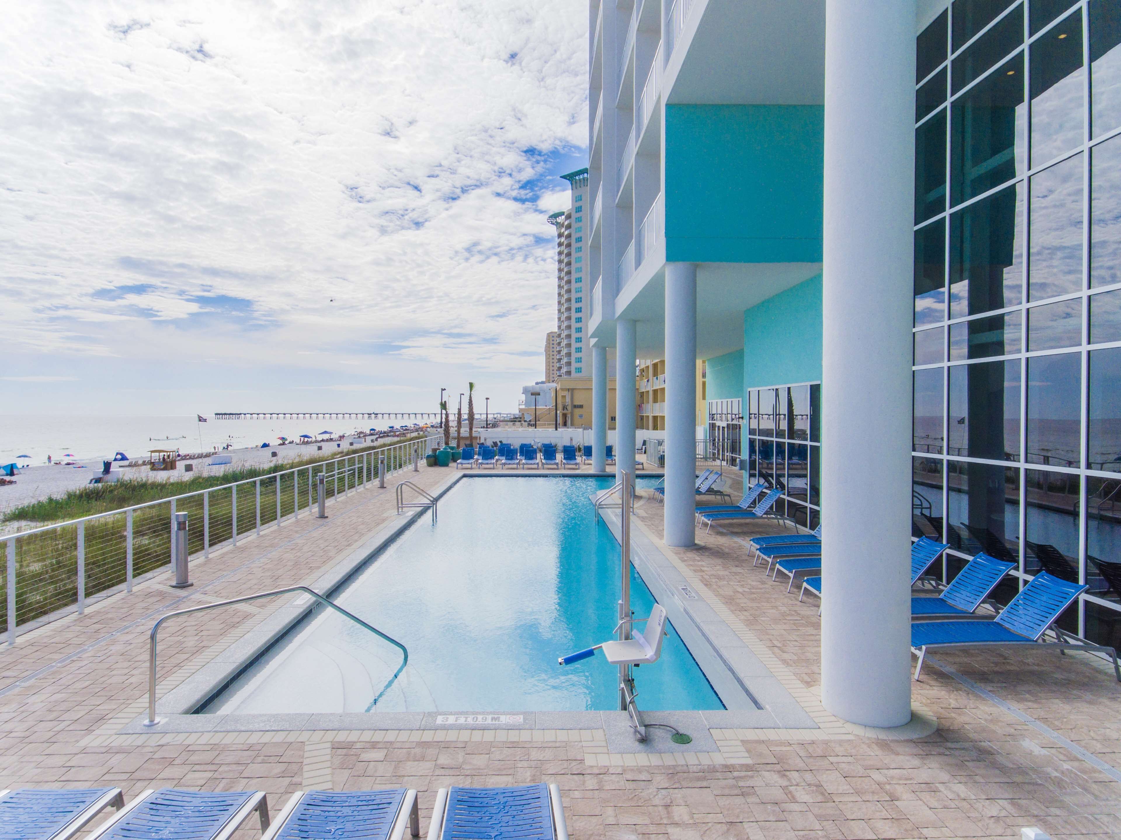 A swimming pool with lounge chairs, a beach view, and a modern building with large windows.