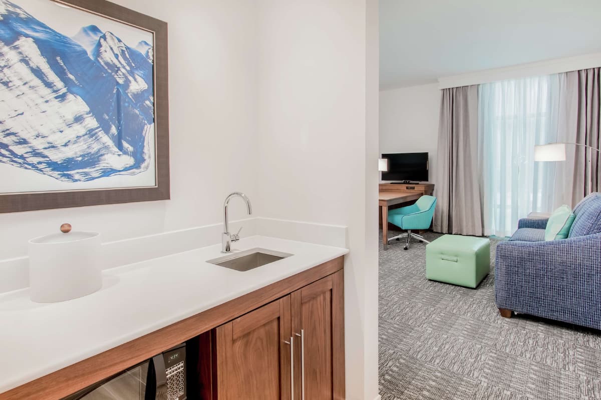 Een moderne hotelkamer met een wit wastafelmeubel, houten kasten, een ingelijste afbeelding, een groene poef, een blauwe bank en een televisie op een houten tafeltje.