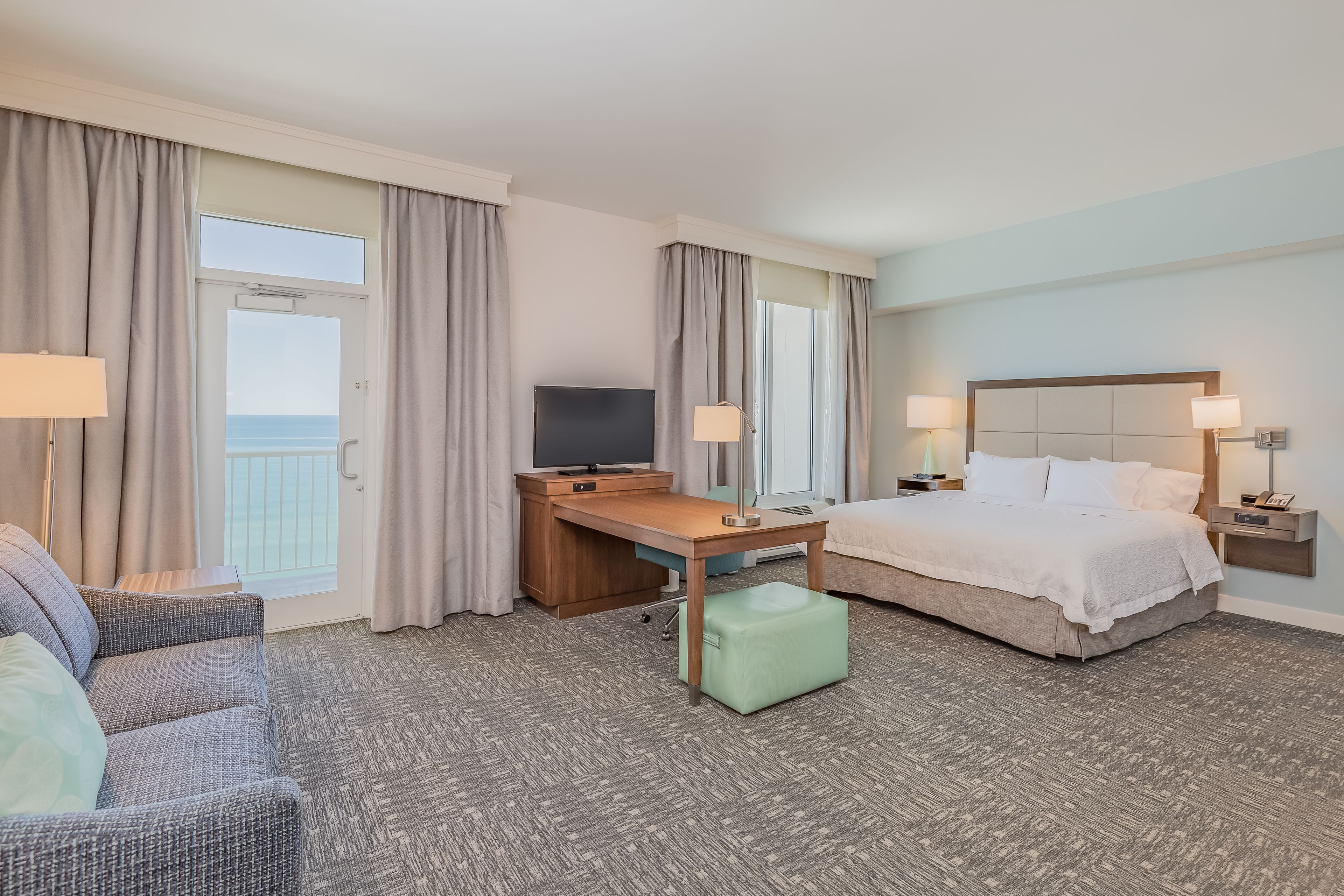 A hotel room with a bed, a desk, a television, and a view of the sea.