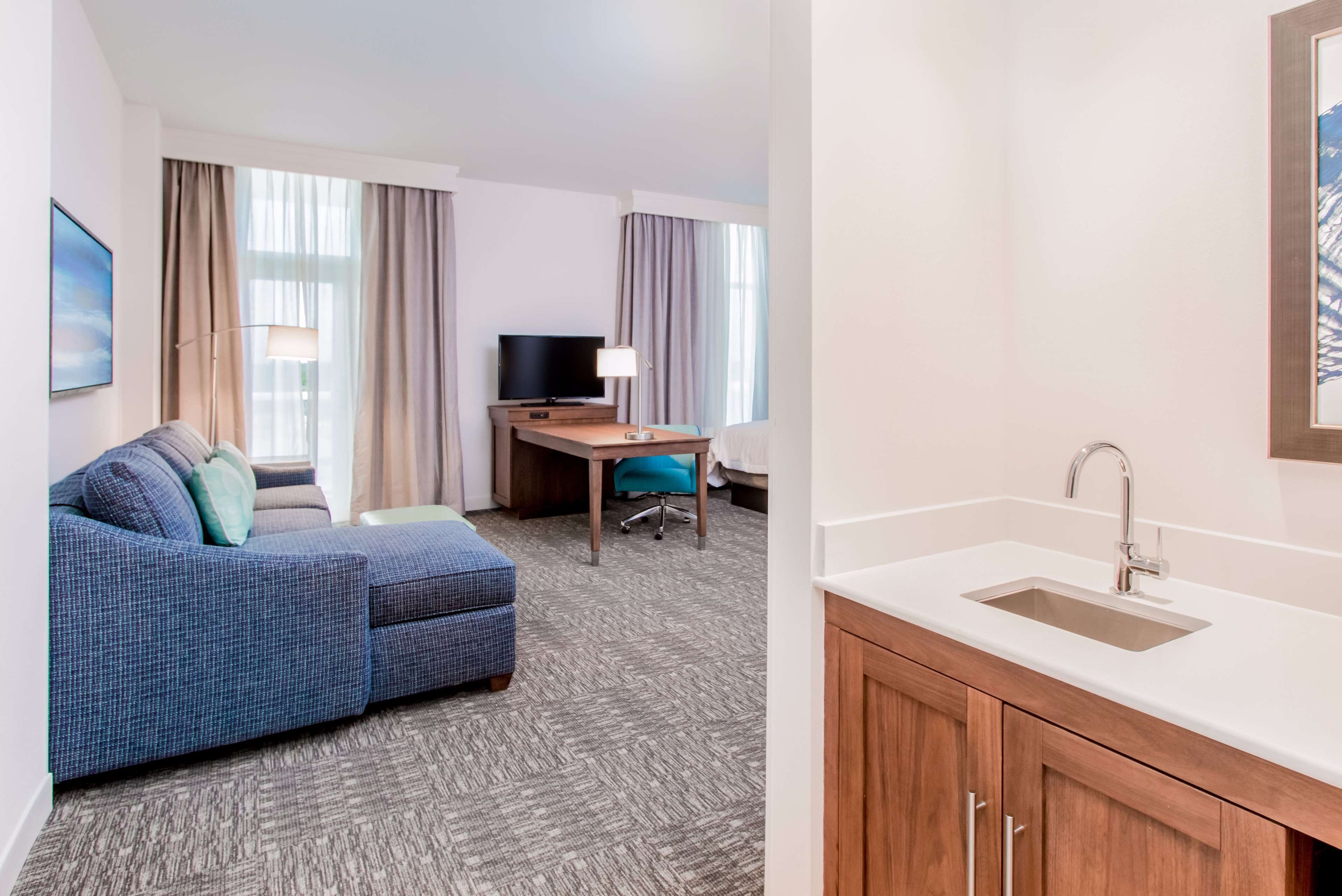 Habitación de hotel con sofá, escritorio con computadora, televisión, lámpara y lavamanos.