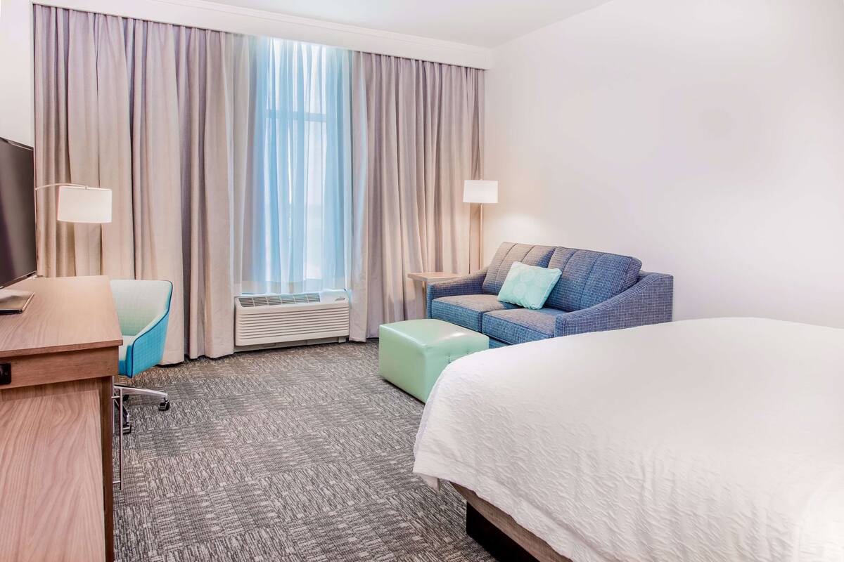 Een hotelkamer met een bed, een bank, een bureau, een stoel, een lamp en een raam met gordijnen.
