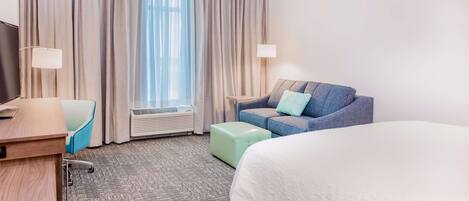 A hotel room with a bed, a sofa, a desk, a chair, a lamp, and a window with curtains.
