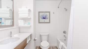 A modern bathroom with a white sink, toilet, and bathtub. There is a framed artwork on the wall and a stack of white towels on a shelf.