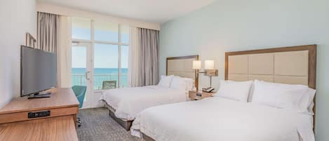 A hotel room with two beds, a desk with a television, and a balcony with a view of the sea.