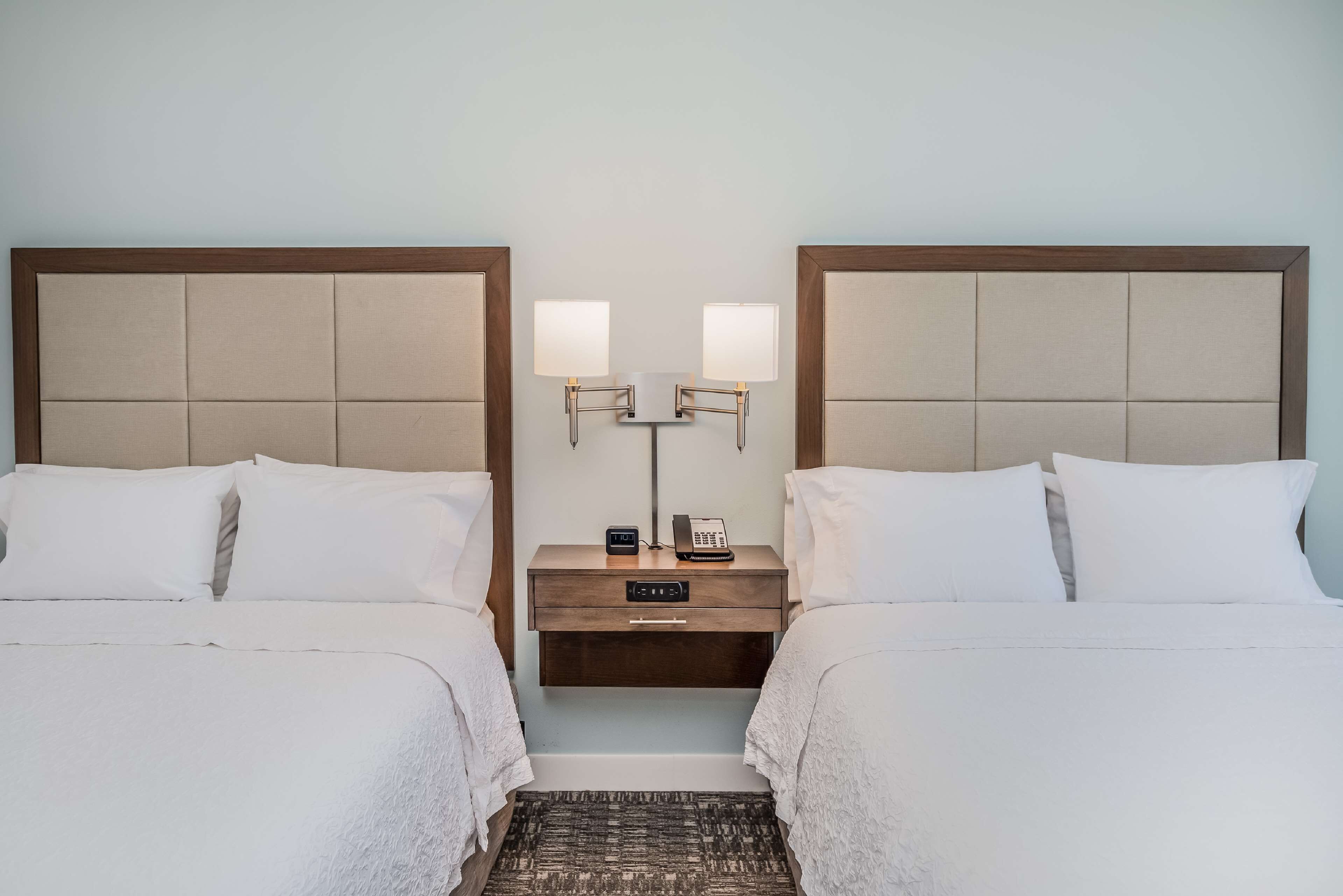 A hotel room with two beds, a nightstand with a lamp, a phone, and a small table.