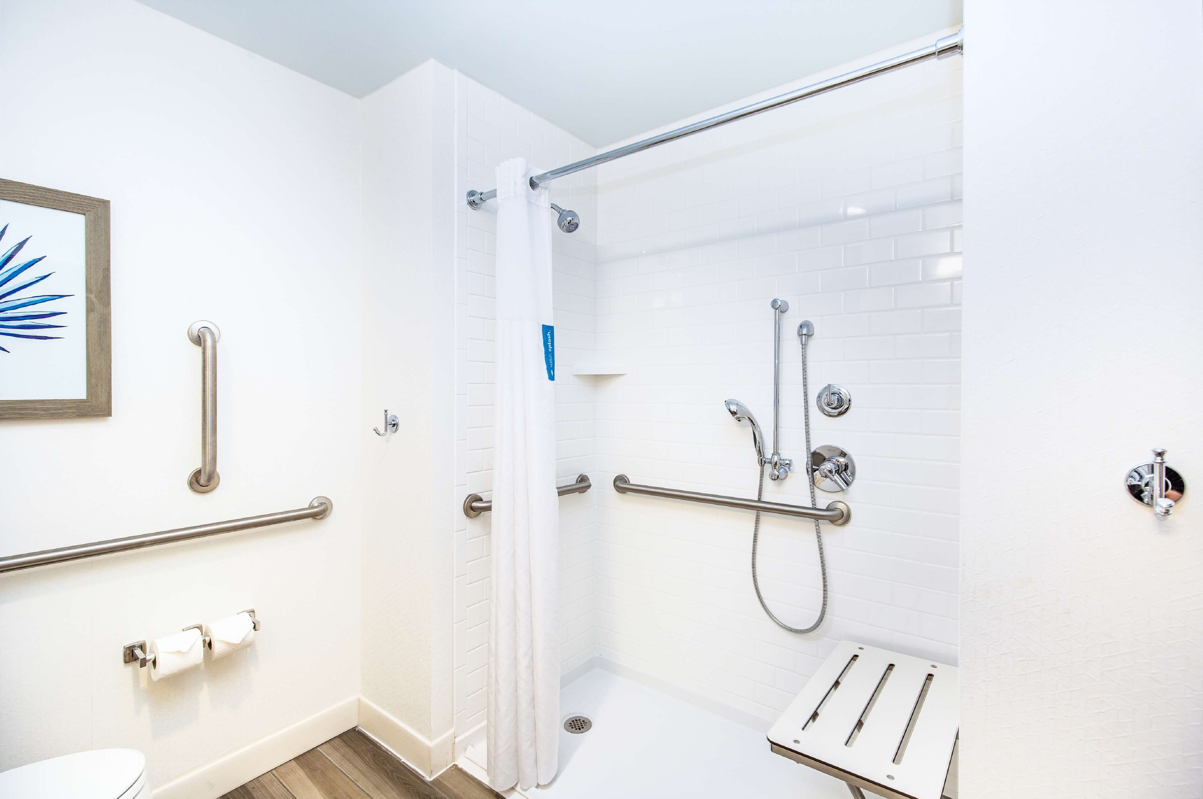 A modern bathroom with white walls, a shower with grab bars, a toilet with a handrail, and a sink with a mirror.