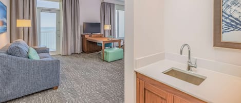 Habitación de hotel con sofá, escritorio, televisión, ventana con cortinas, lavamanos y un cuadro en la pared.
