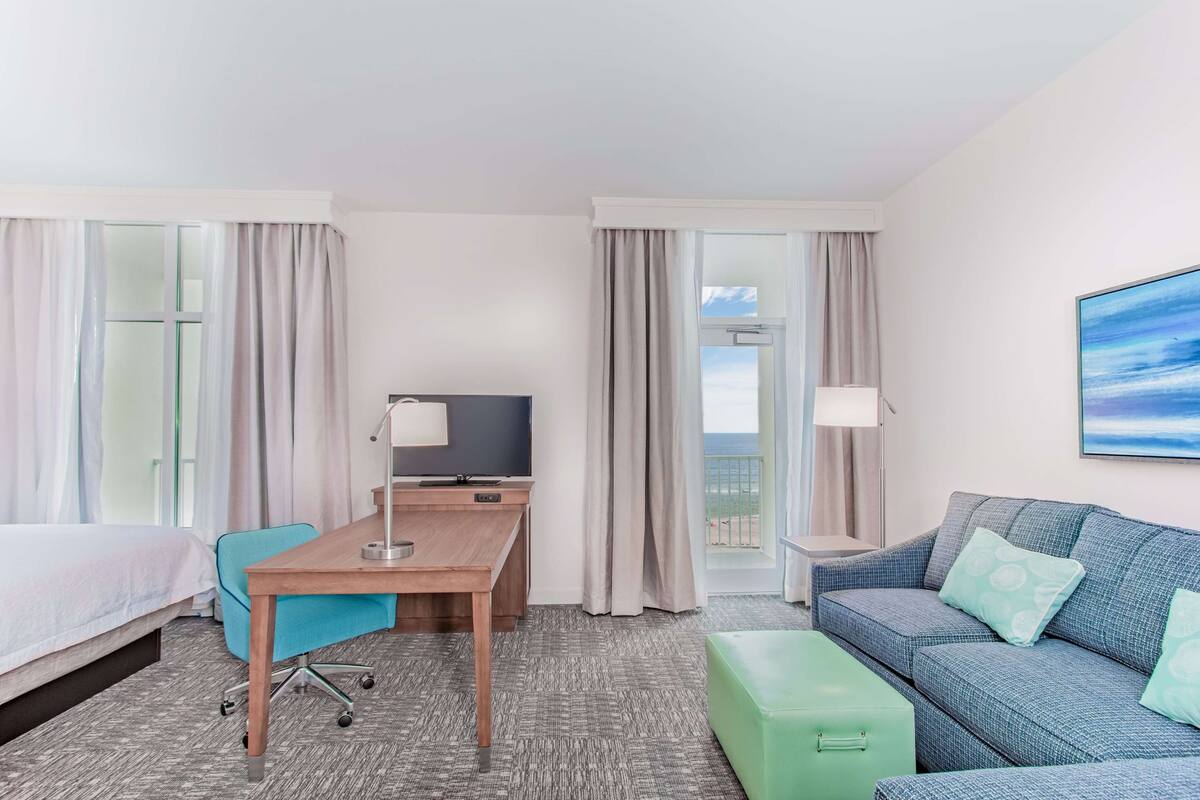 Een hotelkamer met een bed, een bureau met stoel, een bank, een groene poef, een televisie en een balkon met gordijnen.