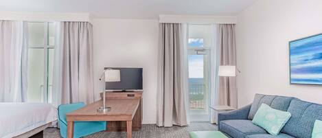 Habitación de hotel con cama, escritorio con silla, sofá, reposapiés verde, televisor y balcón con cortinas.