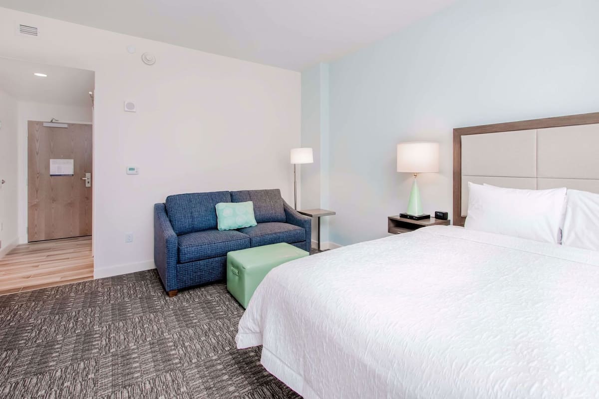 Een hotelkamer met een bed, een slaapbank, een nachtkastje met lamp, een groene poef en een houten deur.