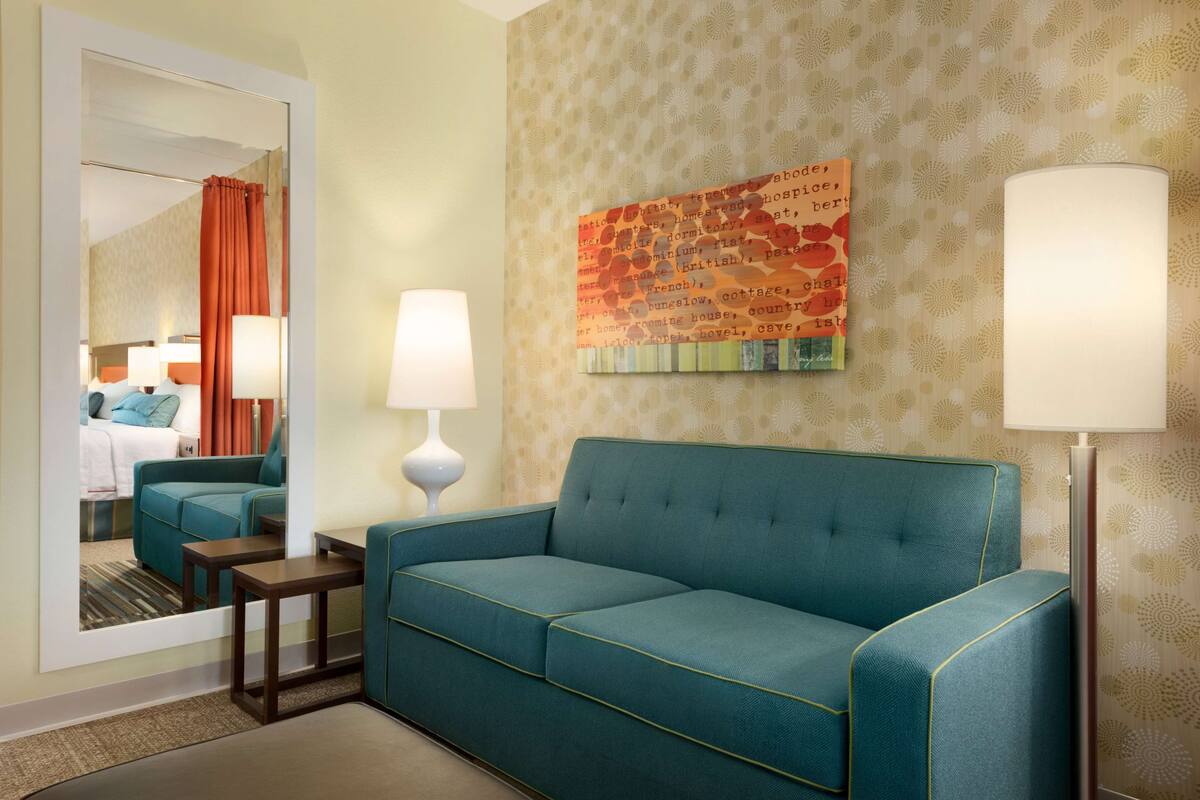 Une chambre d’hôtel avec un canapé de couleur turquoise, un miroir, un mur recouvert d’un papier peint à motifs et un objet décoratif portant une inscription.