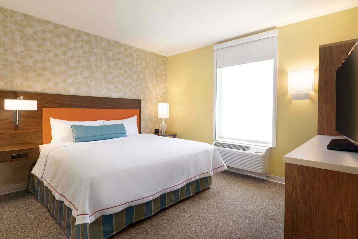 Une chambre d’hôtel avec un grand lit, une table de chevet, un bureau, une télévision et une fenêtre avec des stores.